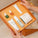 Hands holding an open orange gift box containing Tropic Skincare products, including mini serums, cleanser, moisturiser, toner, and a bamboo face cloth.